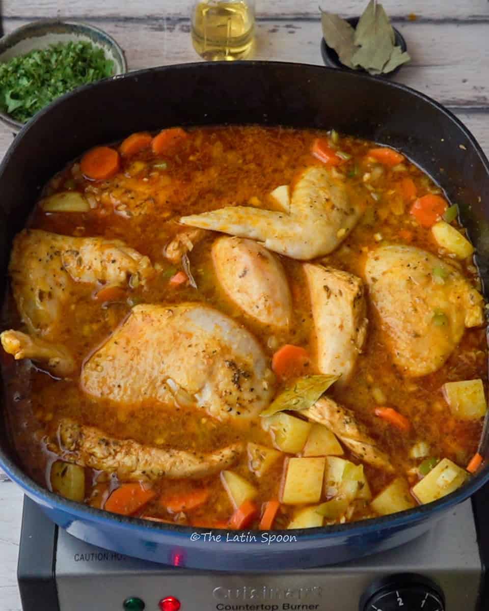 A pot with the mixture of potatoes, carrots, and chicken pieces is shown. In the background, there’s a small cup of cilantro, a container of oil, and a small cup with bay leaves.