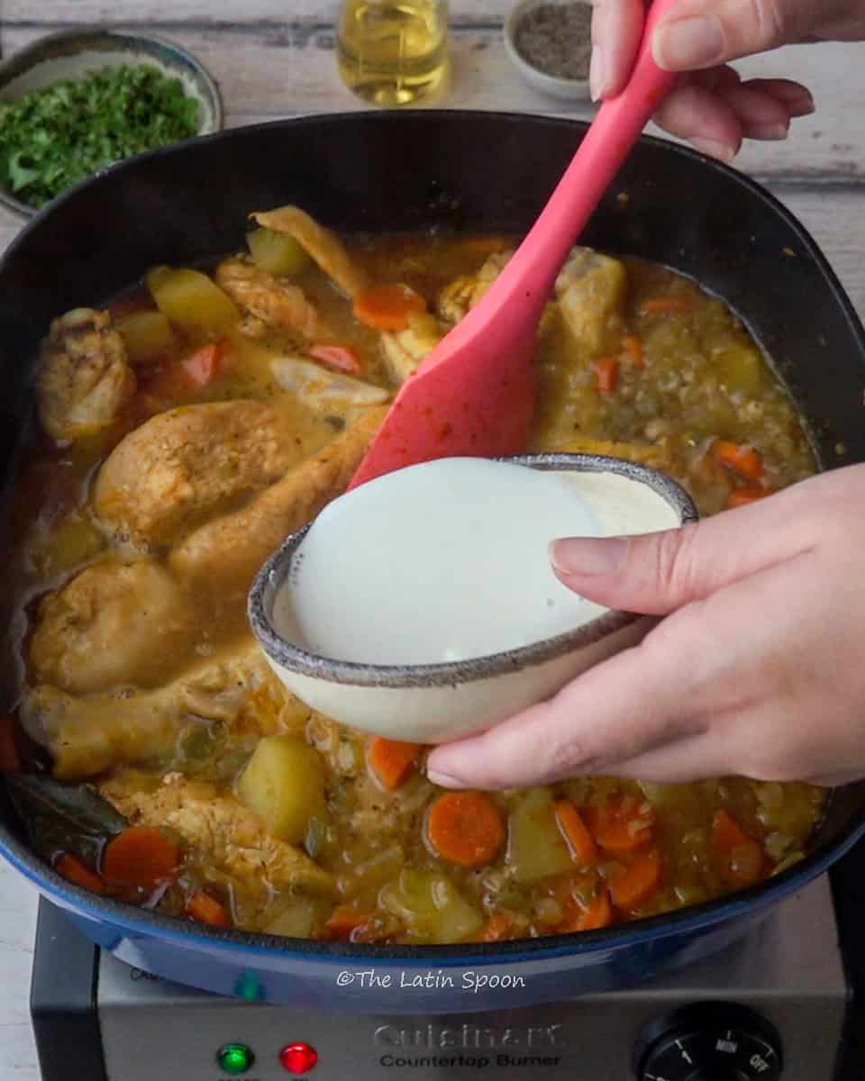 Inside the pot, you can see the mixture of chicken and vegetables, while the left hand holds a small cup of cornstarch, ready to pour it in, and the right hand stirs with a spatula