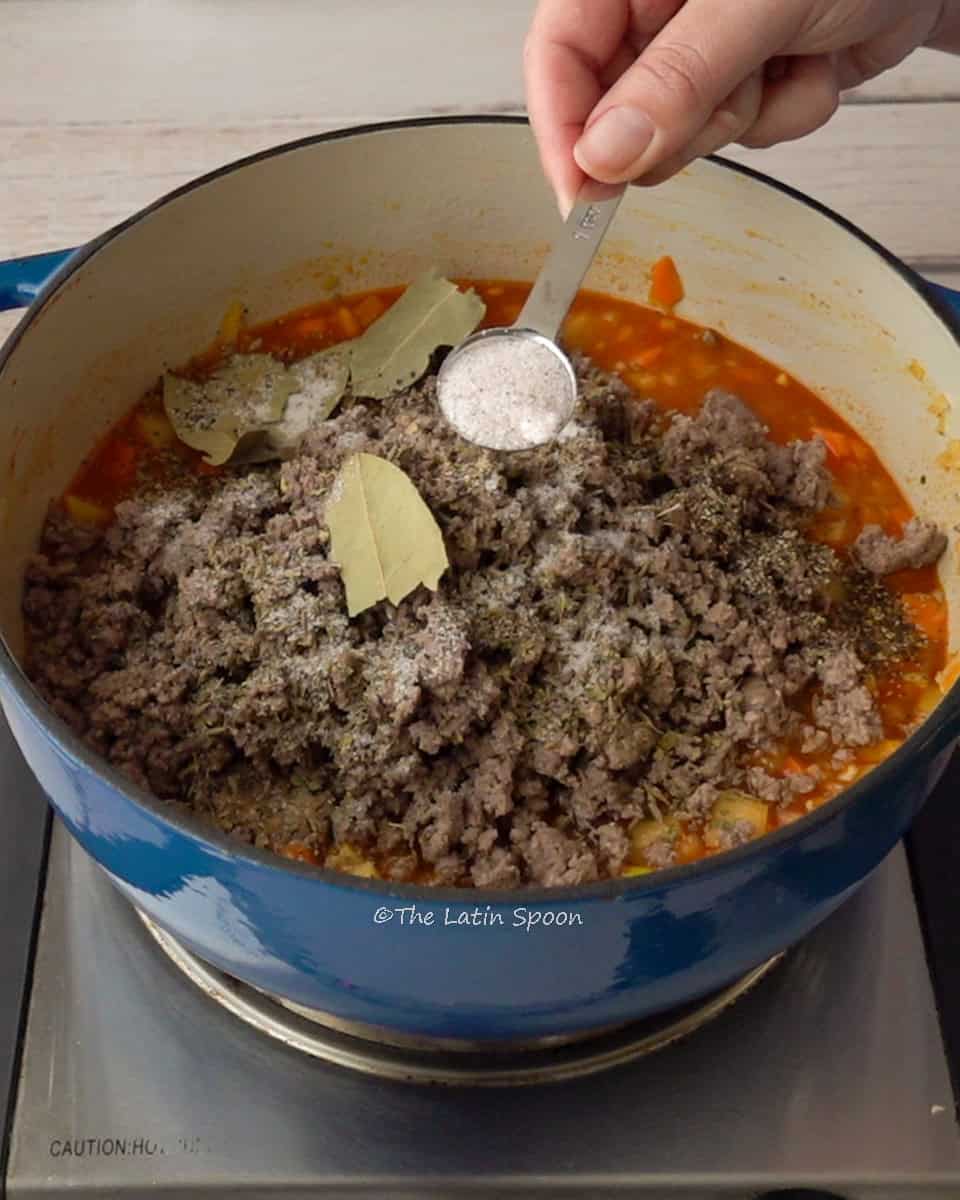 A Dutch oven with the picadillo and ground beef on top with few bay leaves, a hand holding a spoon of salt above as if sprinkling it in.