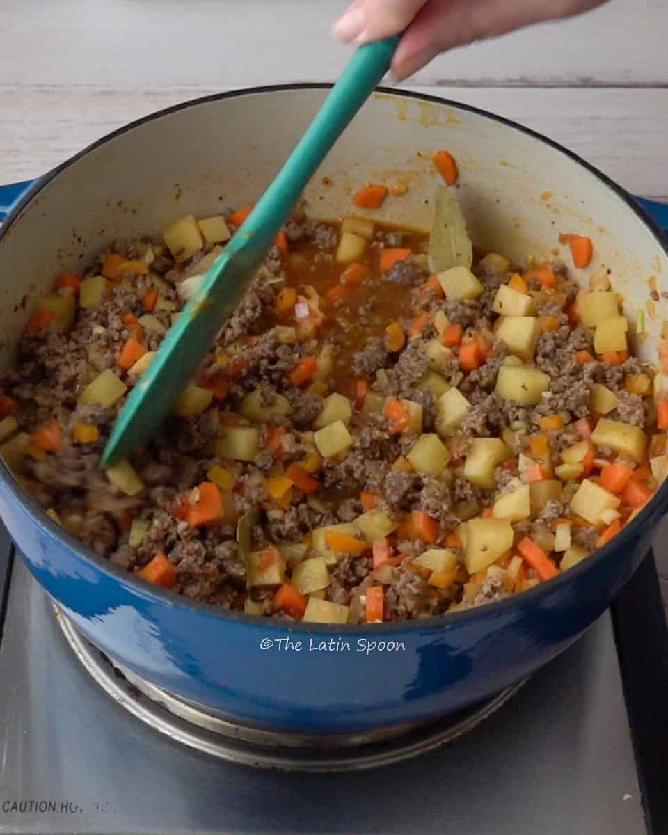 A Dutch oven with ground beef picadillo containing potatoes and carrots, with a hand stirring it using a spatula.