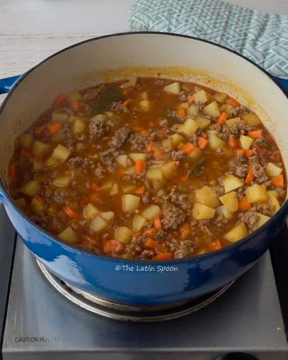 A Dutch oven with ground beef picadillo containing potatoes and carrots, and a kitchen towel in the background.