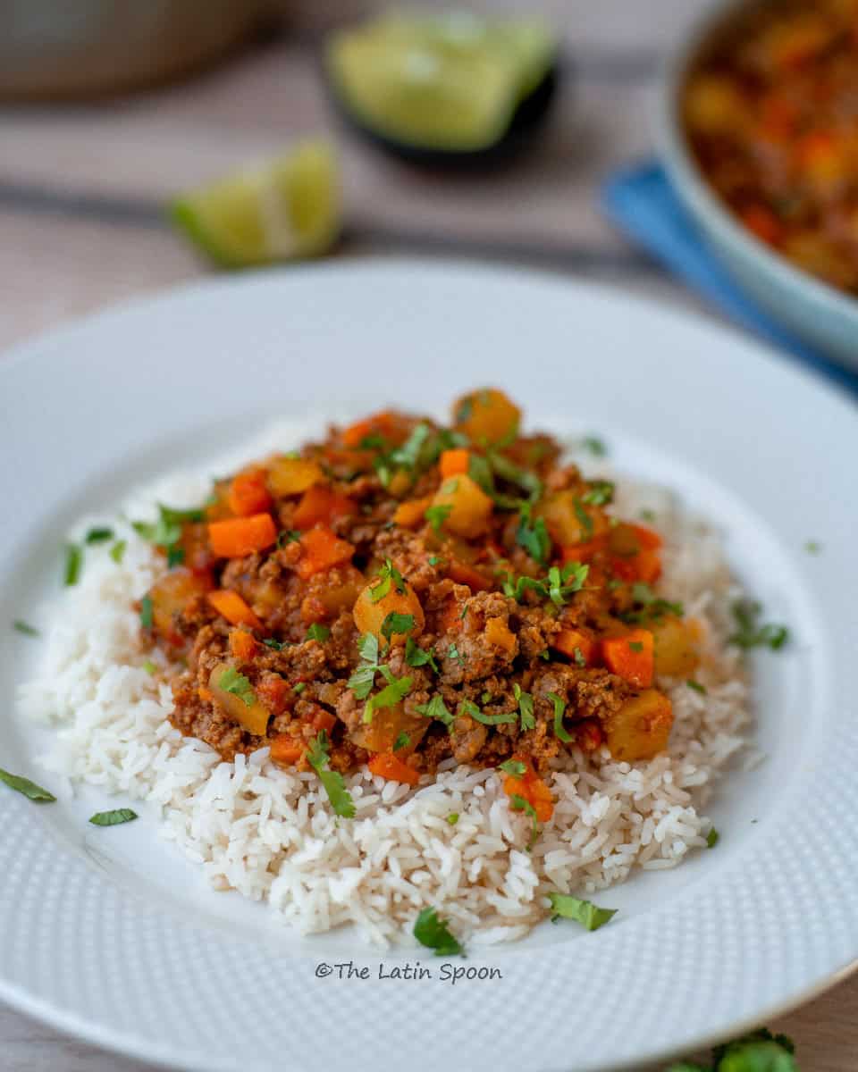 A plate served with a bed of white rice topped with ground beef picadillo with potatoes and carrots.
In the background, a few lemon pieces