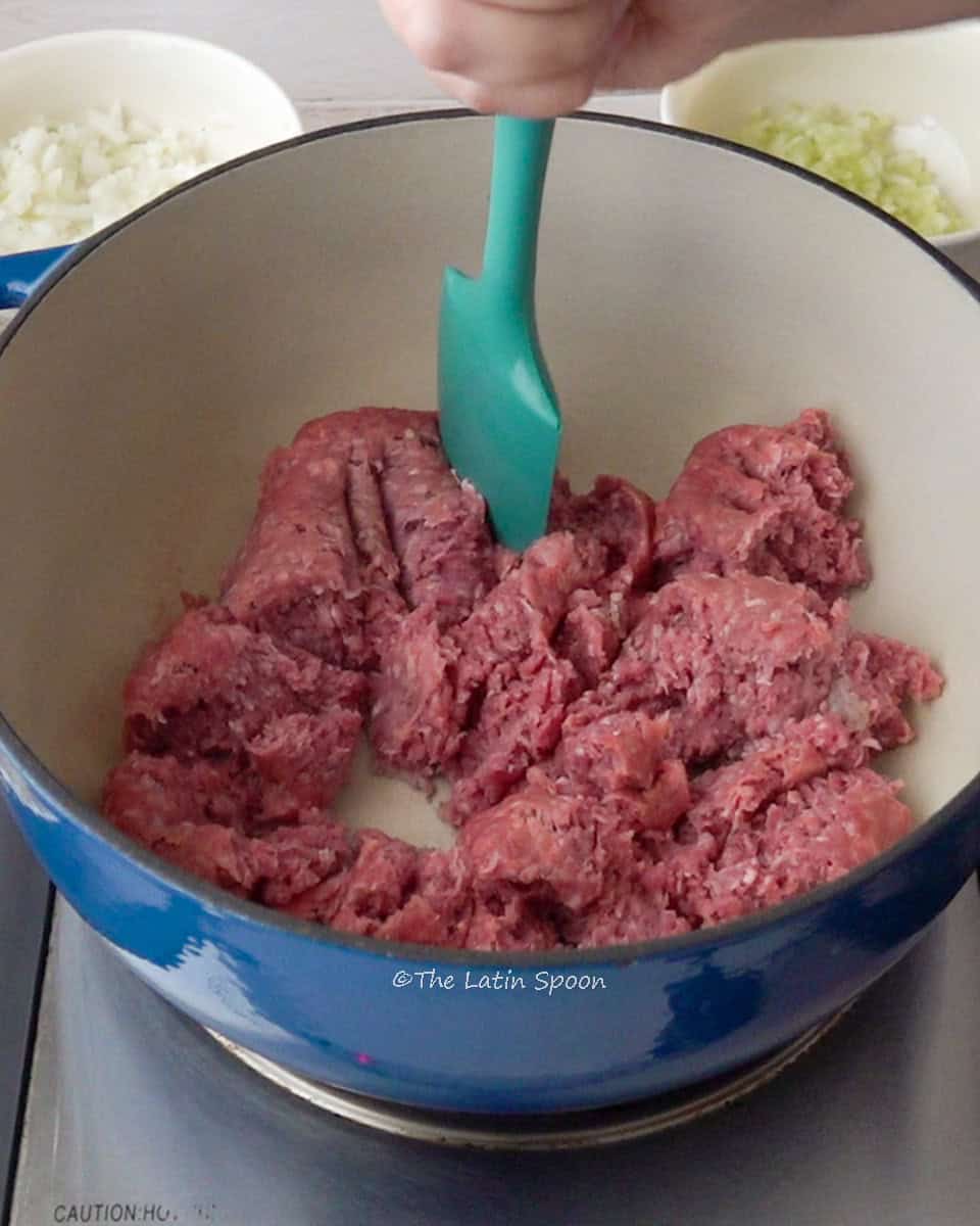 A Dutch oven with raw ground beef inside, a spatula held by a hand stirring it, and small bowls of onion and celery in the background.