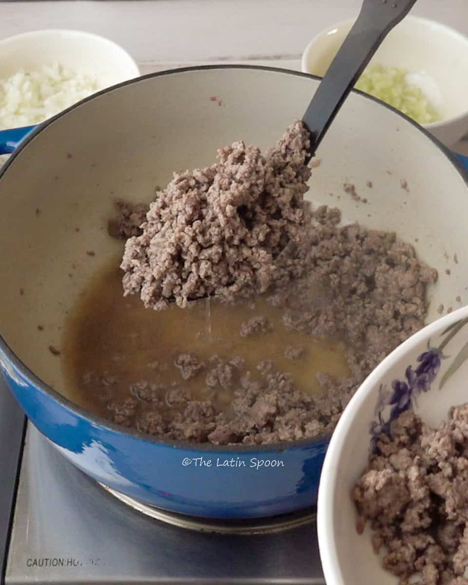 A ladle draining cooked ground beef from the Dutch oven, with more beef remaining at the bottom.
In the background, there are small bowls of onion and celery.