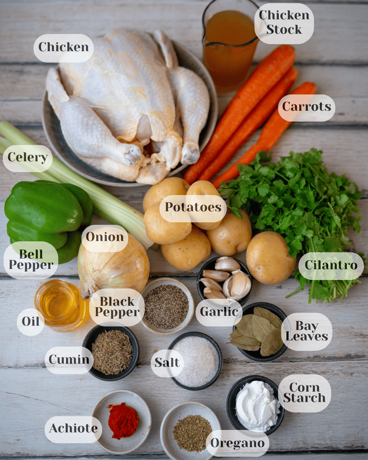 An overhead shot of the ingredients: a whole chicken in a bowl, a small cup of chicken broth, carrots, cilantro, potatoes, garlic, celery, onion, bell pepper, oil, black pepper, bay leaves, salt, cumin, achiote, oregano, and a bit of cornstarch