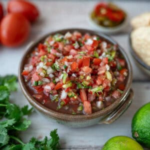A ceramic bowl containing pico de gallo made with tomato, onion, and green chili. In the background, there are tomatoes, a small cup with sliced jalapeños, a small cup with chips, some limes, and cilantro.