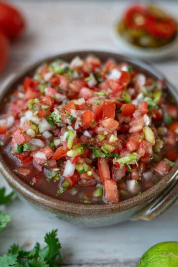 A ceramic bowl containing pico de gallo made with tomato, onion, and green chili. In the background, there are tomatoes, a small cup with sliced jalapeños, a small cup with chips, some limes, and cilantro.