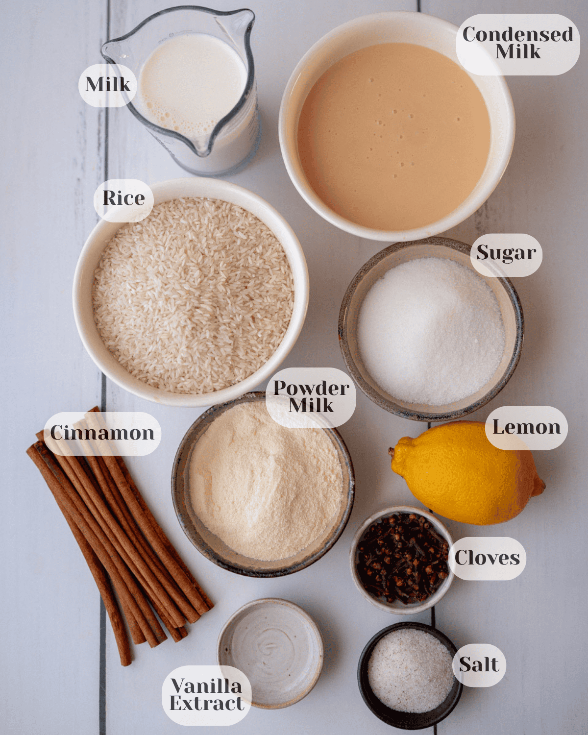 A top-down shot of the ingredients: cup of milk, cup of condensed milk, cup of rice, a small cup of sugar, a small cup of powdered milk, some cinnamon sticks, a small cup of cloves, one lemon, salt, and vanilla extract.

