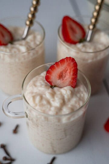 three glass jars filled with rice pudding, each topped with half a strawberry. Two of the jars have a spoon inserted inside. On the countertop around the jars, there are whole cloves scattered, along with a few whole strawberries placed next to them.
