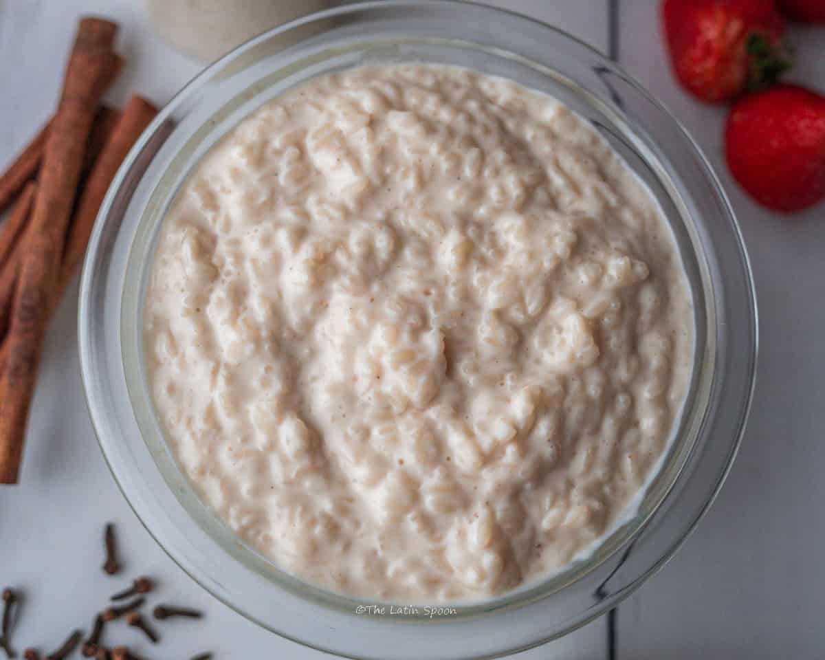 A small cup filled with rice pudding sits in the front. Next to it, there are a few cloves, some strawberries, and a stick of cinnamon. In the background, there are additional cups of rice pudding, each topped with a half strawberry and with spoons placed inside.