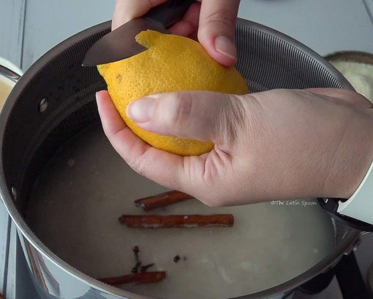 A pot with rice, water, cloves, and cinnamon sticks is visible in the back. One hand holds a lemon, and the other hand cuts the peel with a knife