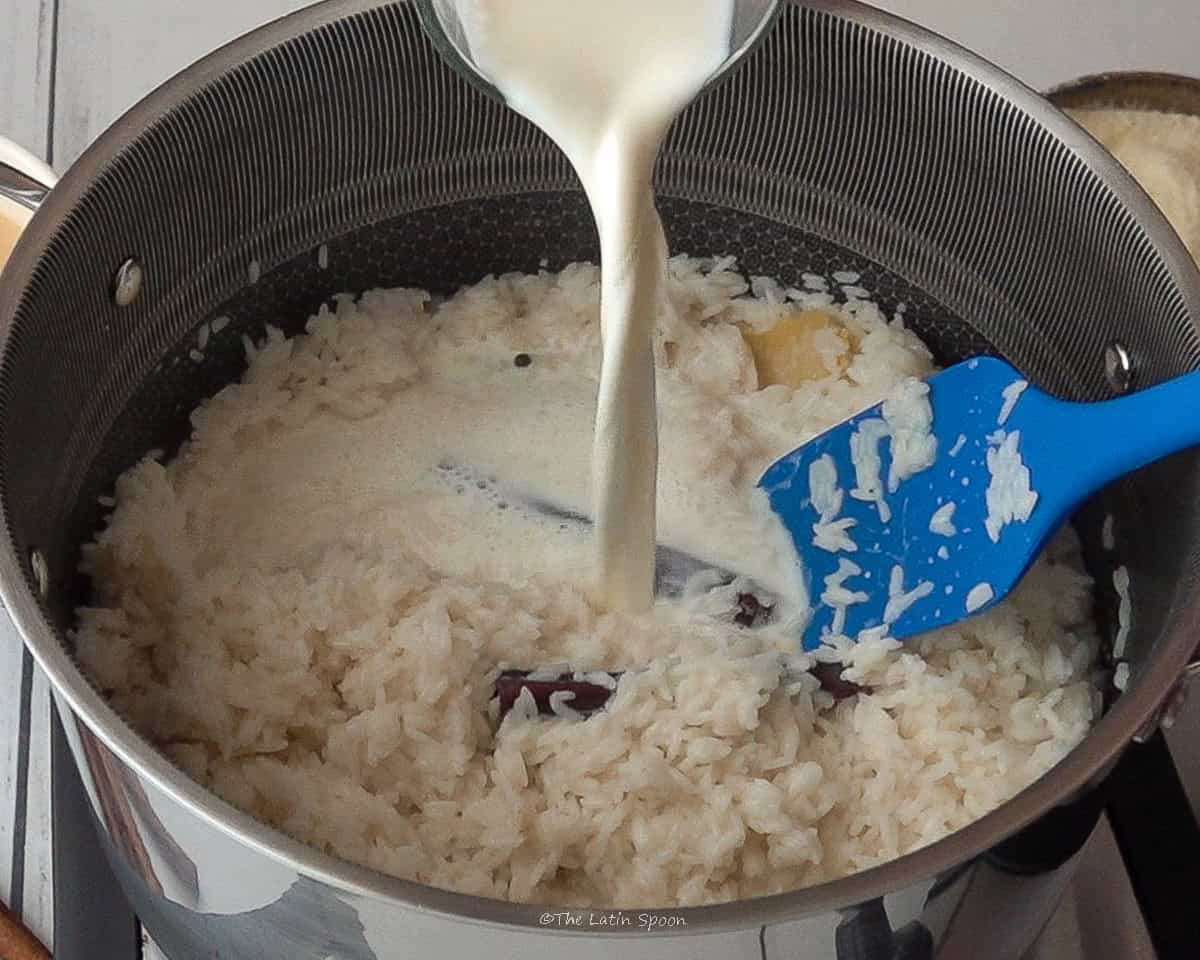 A pitcher of milk being poured into a pot containing rice, cinnamon sticks, cloves, and small pieces of lemon peel.