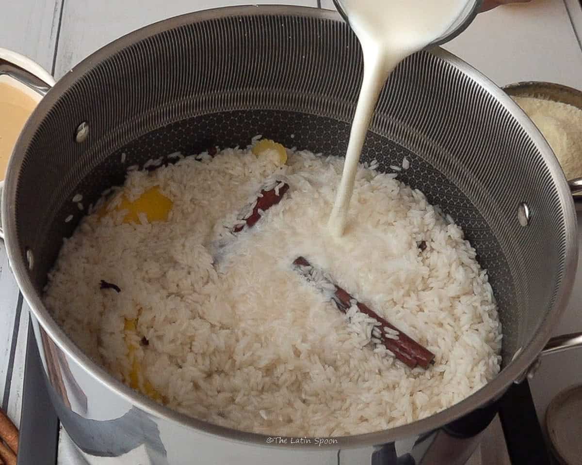 A pitcher of milk being poured into a pot containing rice, cinnamon sticks, cloves, and small pieces of lemon peel.