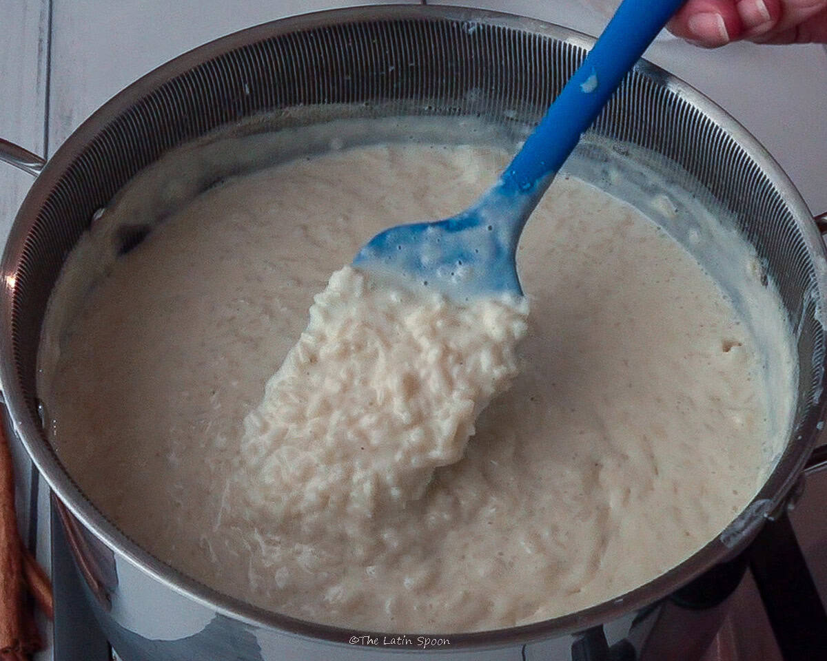 A hand holds a spatula inside the pot of rice pudding, lifting a small portion of the mixture. In the background, slightly out of focus, there is a small cup filled with cloves.