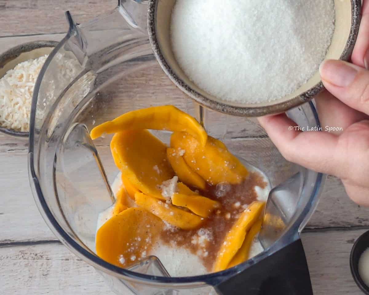 A blender cup containing mango pieces, milk, and vanilla extract, with a hand holding a cup of sugar above it.