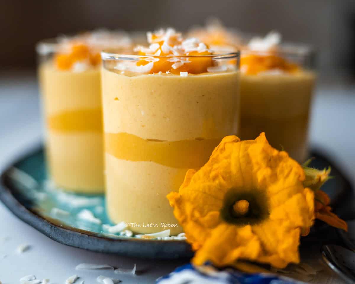 A serving platter with small glass cups filled with mango mousse, topped with mango pieces and coconut flakes, with a decorative flower placed on the platter.