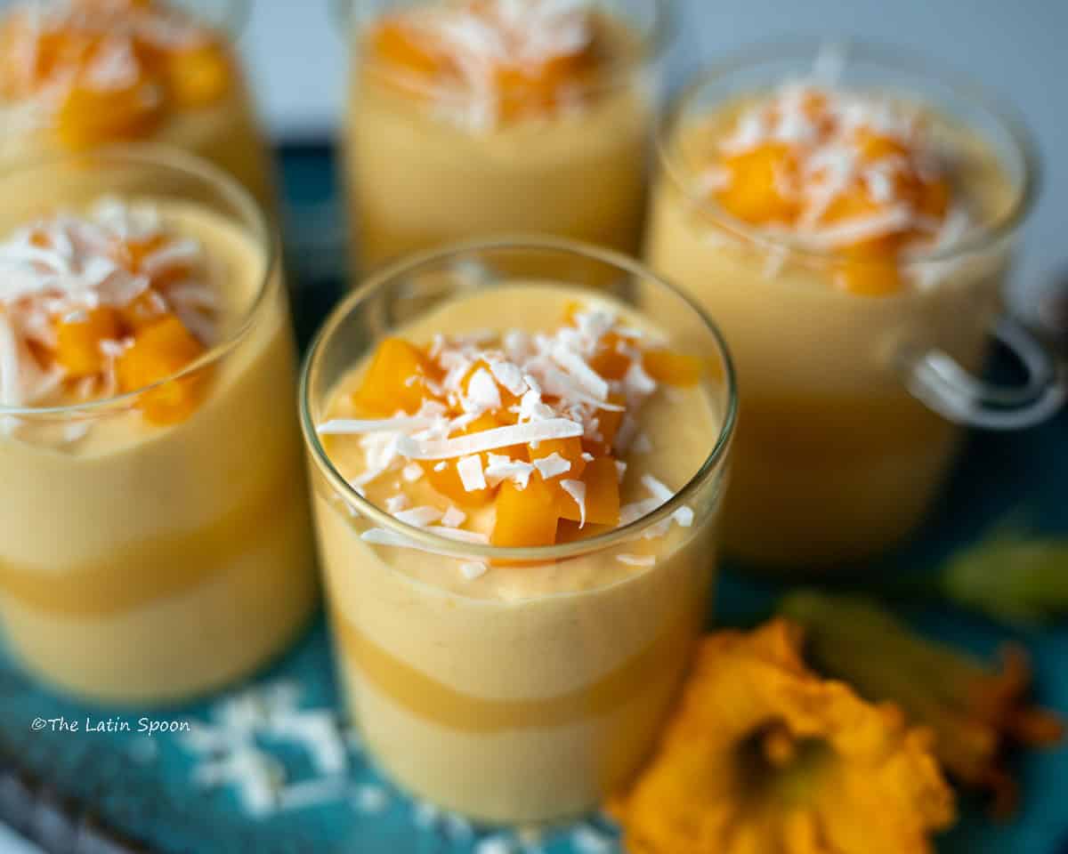 Top view of a decorative platter holding small glass cups filled with mango mousse, topped with mango pieces and coconut flakes, with an edible flower placed on the side for decoration.