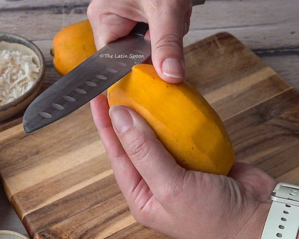 One hand holding a mango while the other uses a knife to peel it, with a cutting board visible in the background.