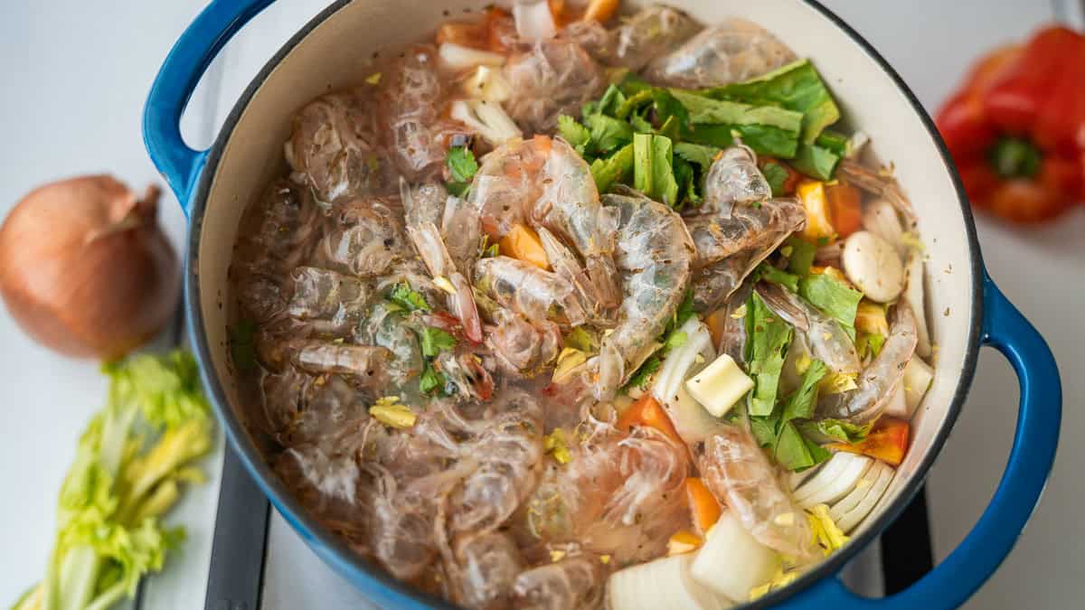 A pot containing shrimp shells and vegetables. In the background, an onion, a celery stalk, and a bell pepper.