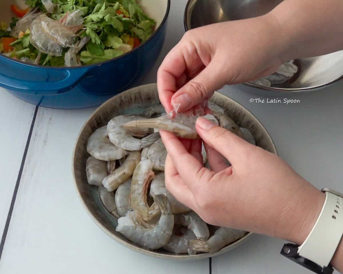 There is a pot with vegetables and some shrimp shells, a bowl with peeled shrimp, and a bowl with unpeeled shrimp in the front. Above the bowl, two hands are removing the shell from a shrimp.