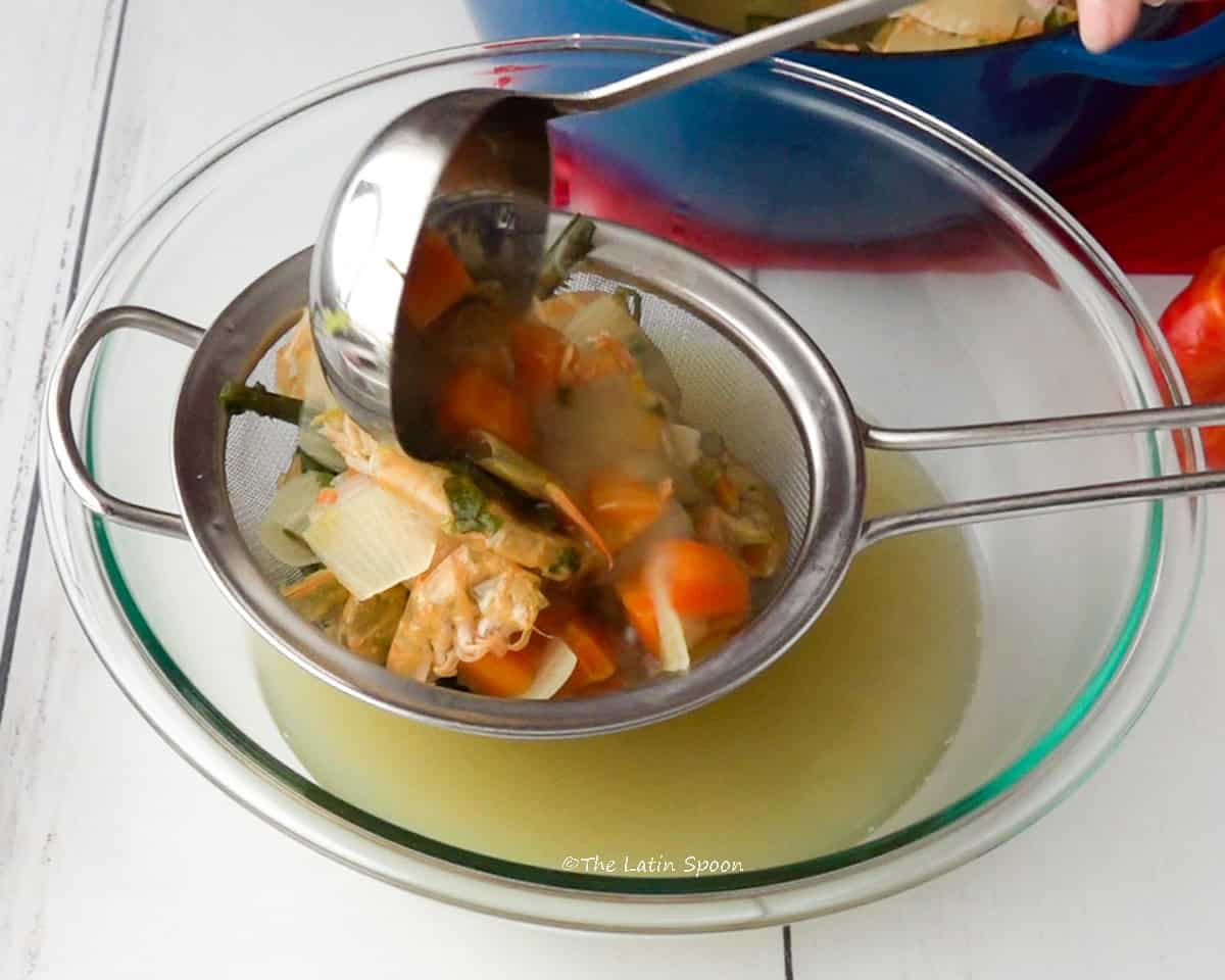 A bowl with a strainer on top, while the vegetables and shrimp shells are being poured through it to strain the broth