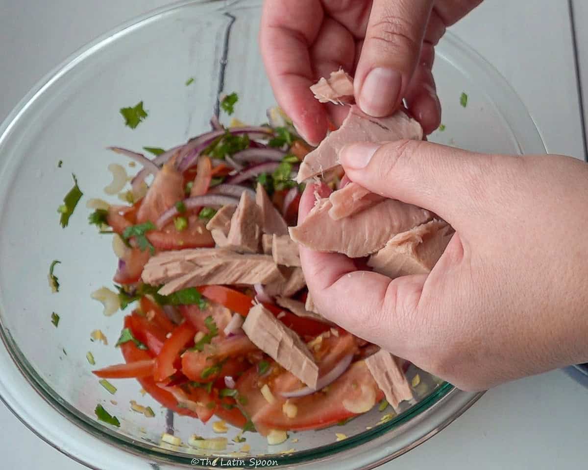 A glass bowl in the background with two hands separating tuna chunks into smaller pieces.