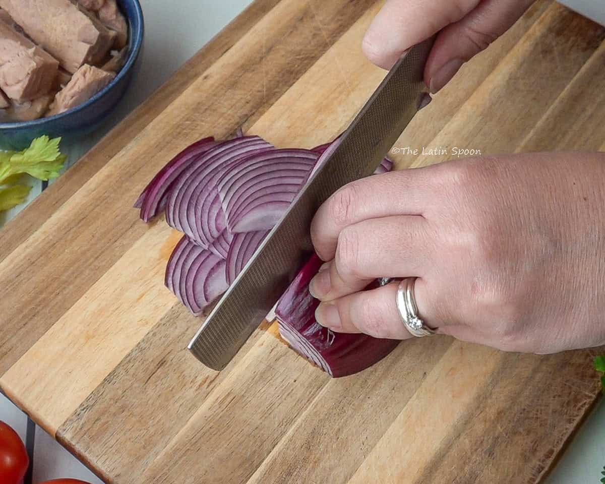 A wooden cutting board with red onion being sliced into long strips, with a bowl of tuna chunks in the background.