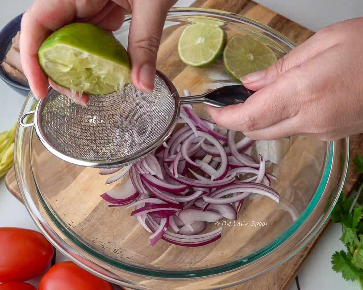 A glass bowl with red onion strips inside, a strainer on top, and a right hand squeezing lemon juice over the onions.