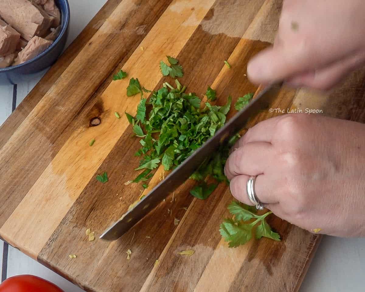 A wooden cutting board with two hands finely chopping cilantro.