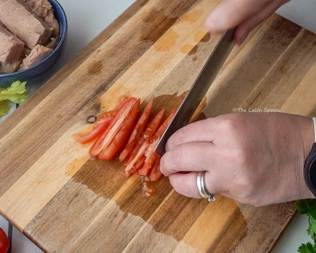 A wooden cutting board with two hands slicing tomatoes into long strips lengthwise.