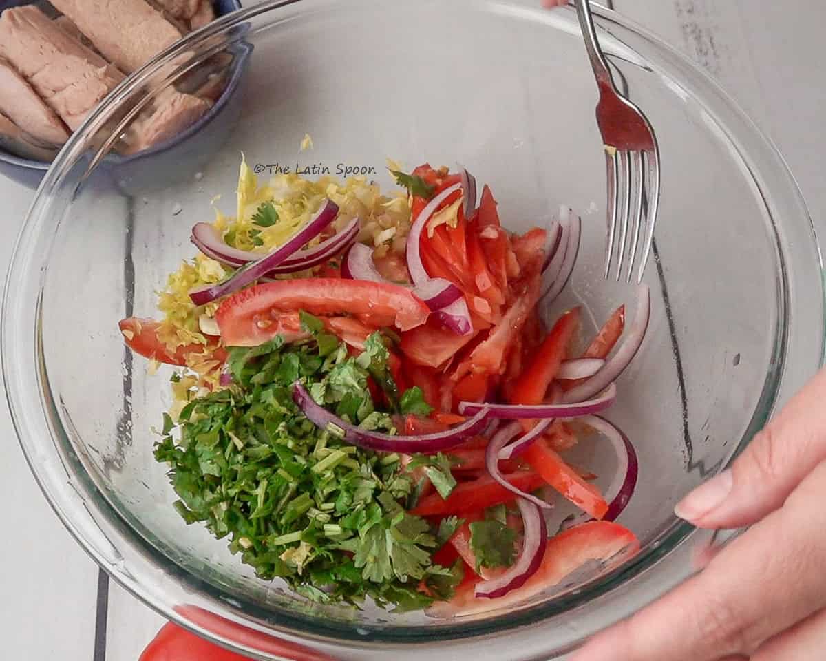 A glass bowl with all the chopped ingredients, with a fork approaching the bowl to begin mixing.