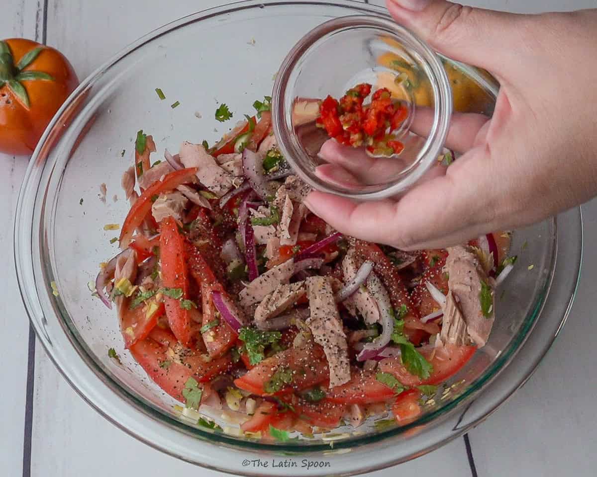 A glass bowl holding the tuna salad while a small cup above it adds minced jalapeño to the salad.