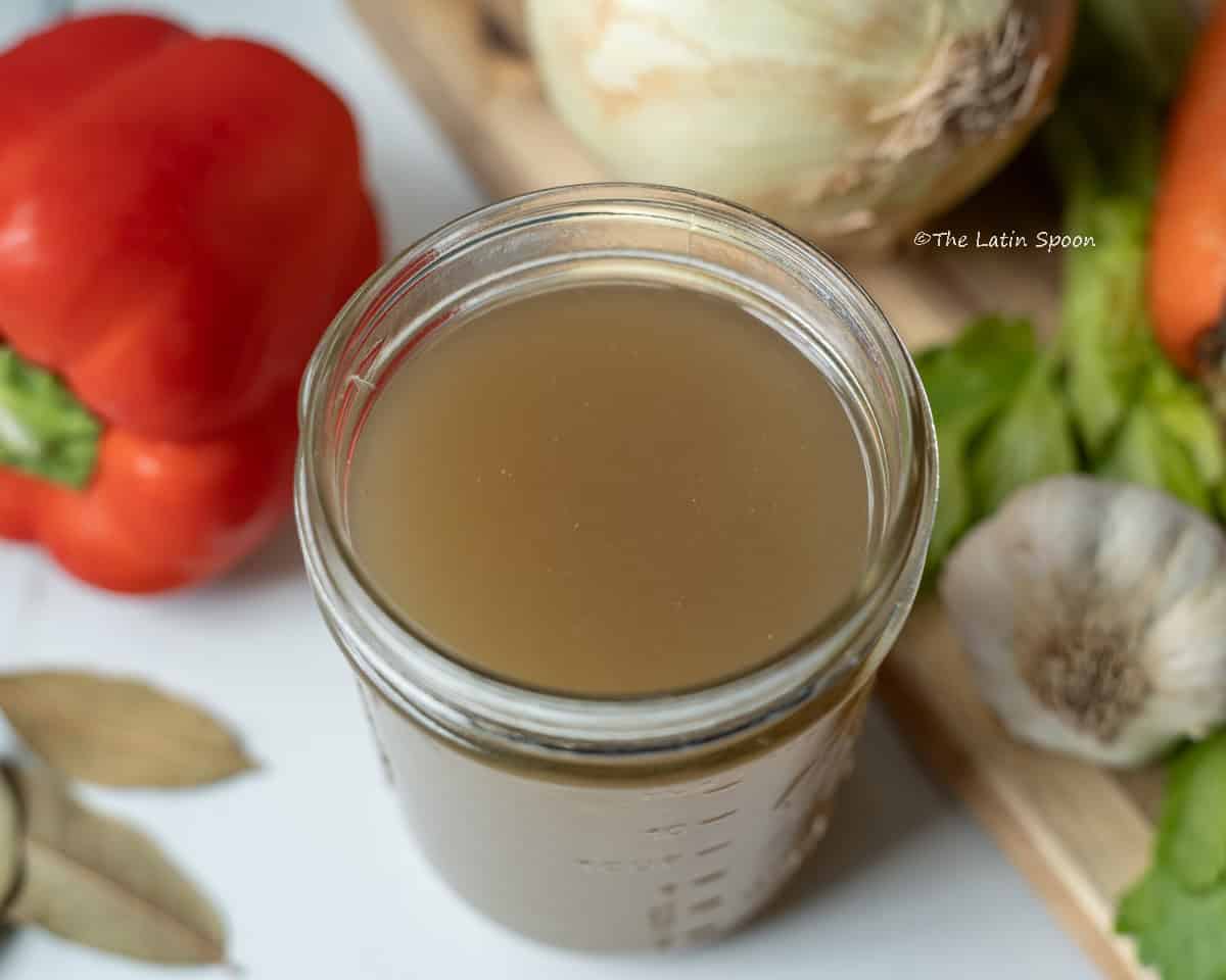 Jar of homemade chicken broth surrounded by carrots, celery, onions, garlic, bay leaves, and bell peppers.