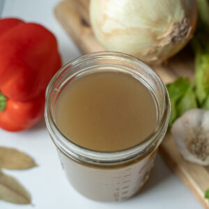 Jar of homemade chicken broth surrounded by carrots, celery, onions, garlic, bay leaves, and bell peppers.