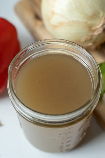 Jar of homemade chicken broth surrounded by carrots, celery, onions, garlic, bay leaves, and bell peppers.