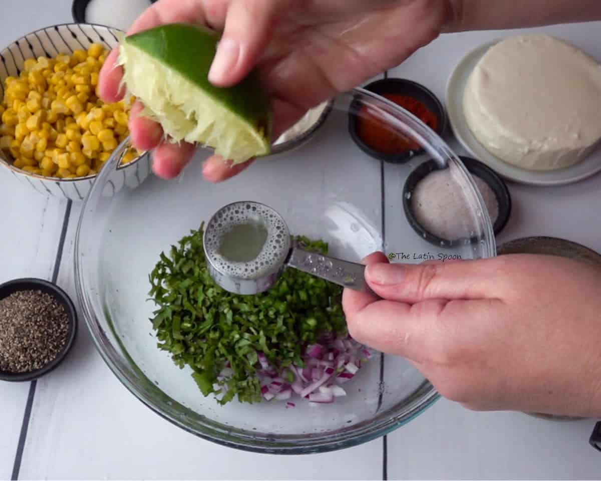A bowl with red onion, culantro, and cilantro. Above it, two hands can be seen: one holding a measuring cup and the other squeezing half a lemon to add the juice into the cup. Next to the bowl, there are sweet corn, cheese, and seasonings in small bowls.