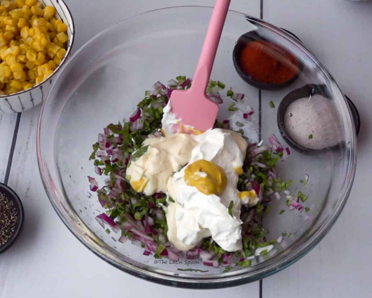 A bowl with red onion, culantro, cilantro, jalapeño pepper, sour cream, mayonnaise, and mustard, with a spatula inside. Next to the bowl, there is sweet corn in a cup, paprika, and salt.