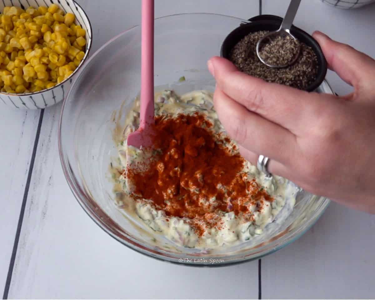 A bowl with the dip mixture and paprika sprinkled on top. One hand is holding a small bowl of black pepper, while the other hand holds a measuring spoon with black pepper. Next to the bowl, there is a small bowl of sweet corn.