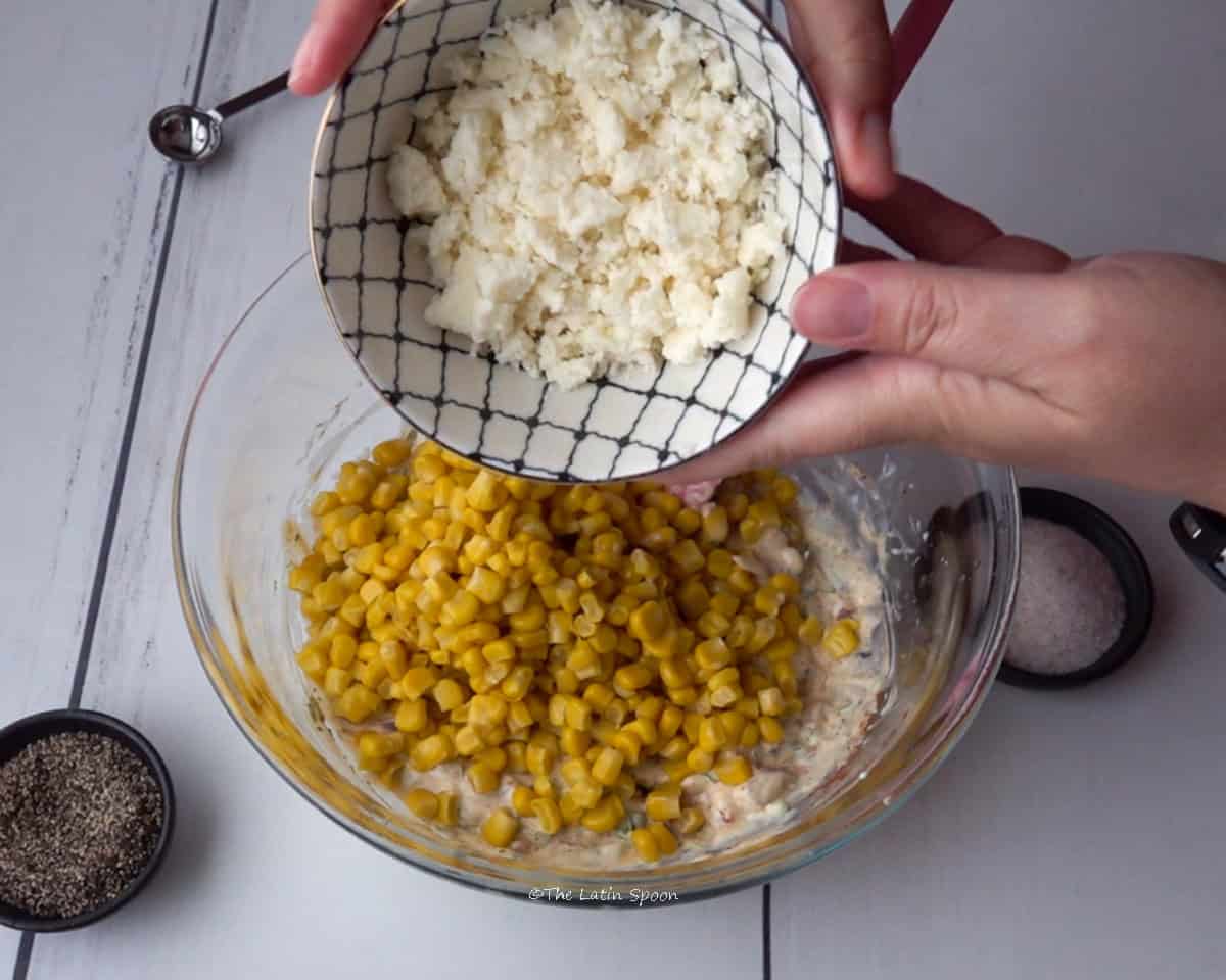 A bowl with the dip mixture and corn on top. Above the bowl, two hands are holding a cup of fresh cheese. On the side, there are small bowls of salt and black pepper.