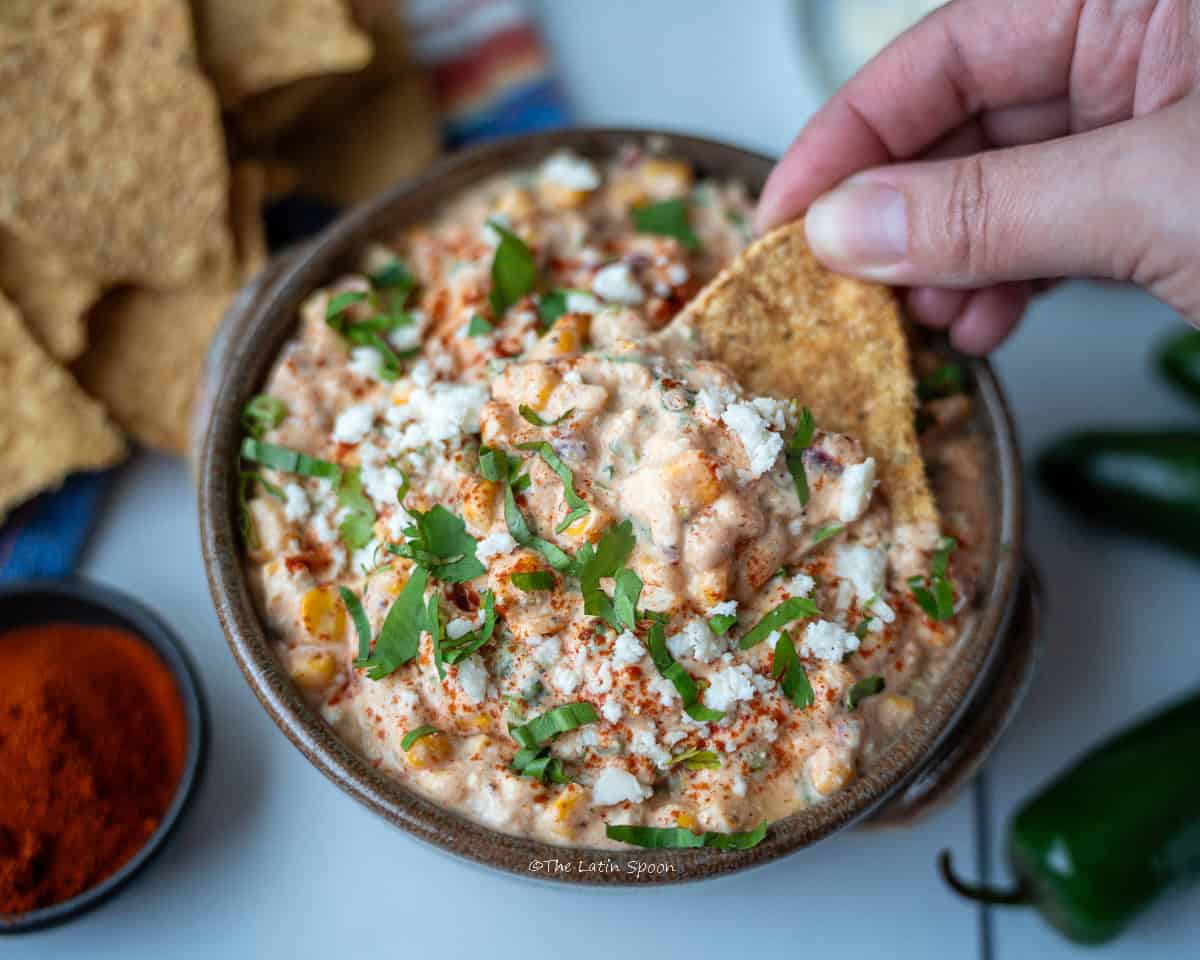 A deep bowl of corn dip, topped with paprika, chopped cilantro, and cheese. The photo is taken from above; on the side, there are toasted tortilla chips, a small bowl of paprika, and whole jalapeño peppers, while a hand holds a chip with dip.