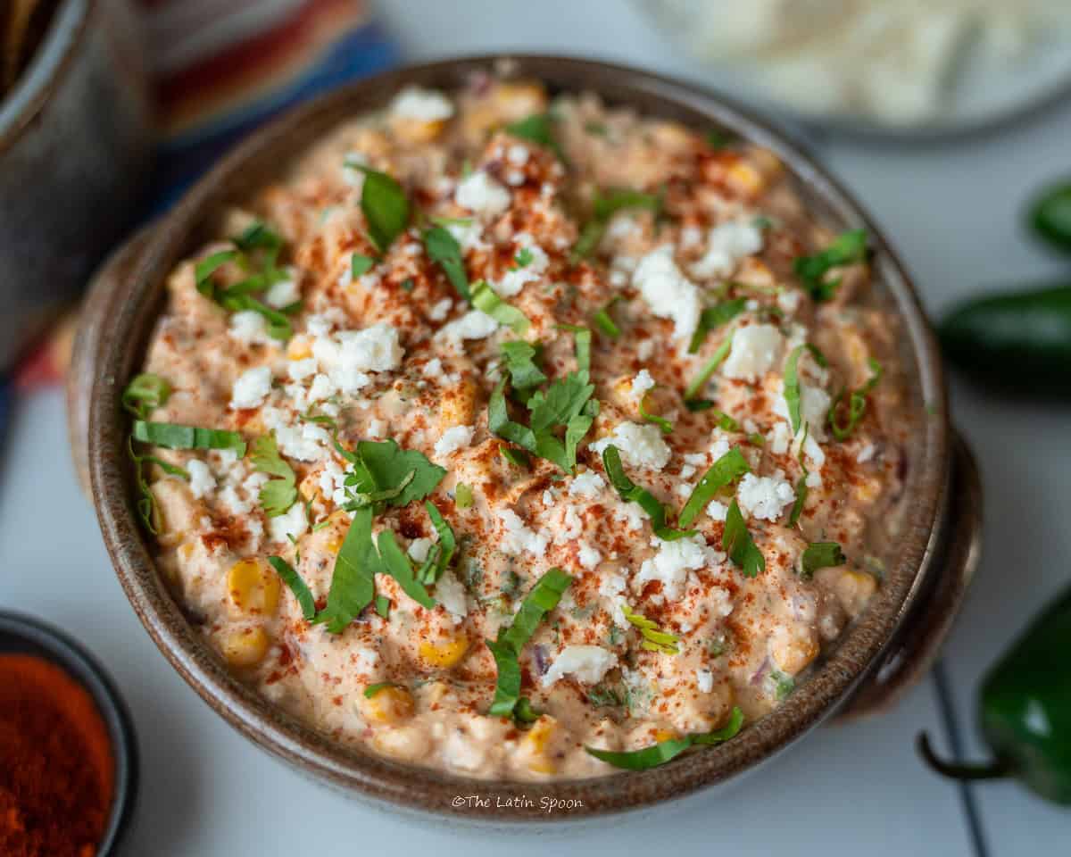 A deep bowl of corn dip, topped with paprika, chopped cilantro, and cheese. The photo is taken from above, with toasted tortilla chips and a small bowl of paprika on the side.
