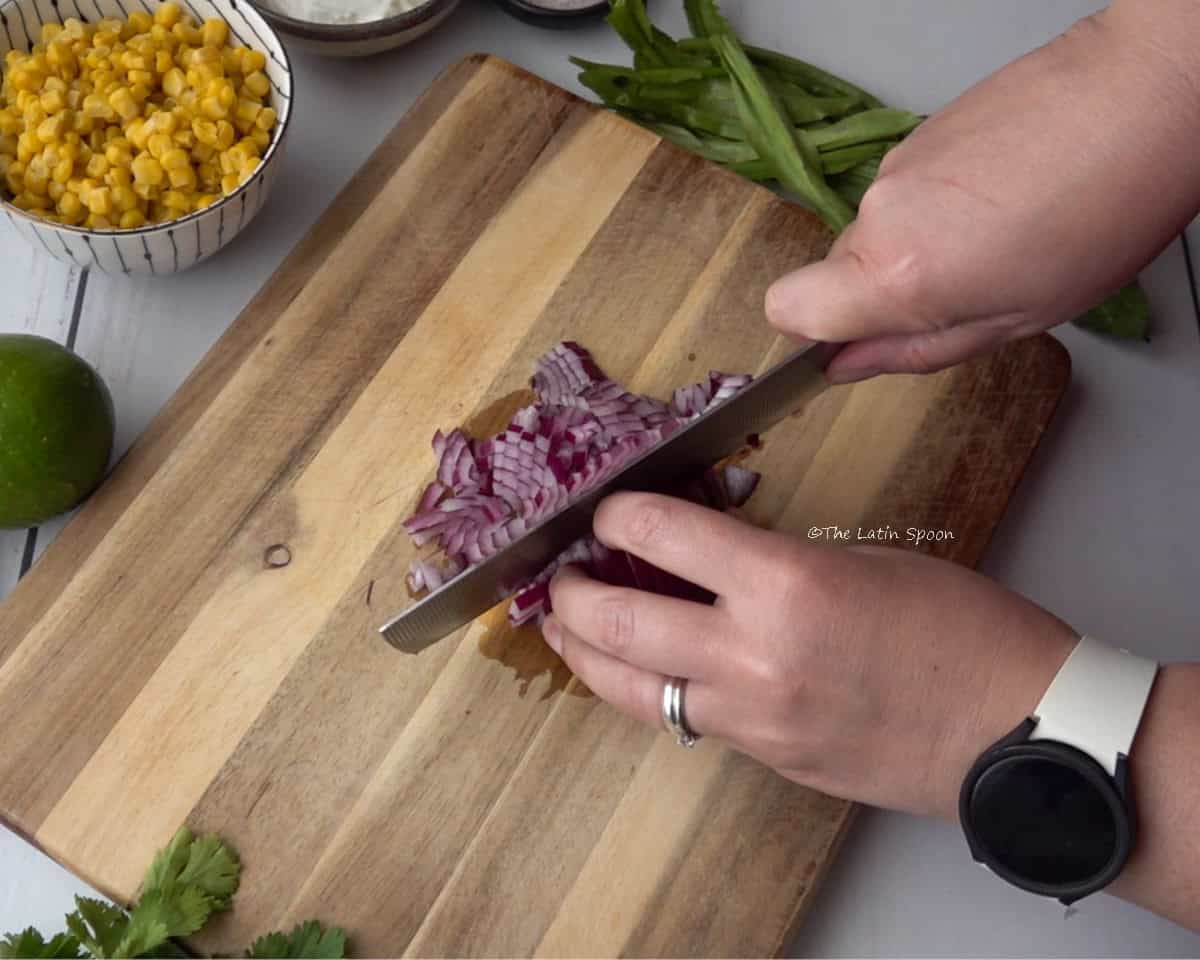 A cutting board with a red onion being sliced. One hand is holding the onion while the other holds the knife. Around it, there is a small bowl of sweet corn, a bunch of cilantro, and a lemon.