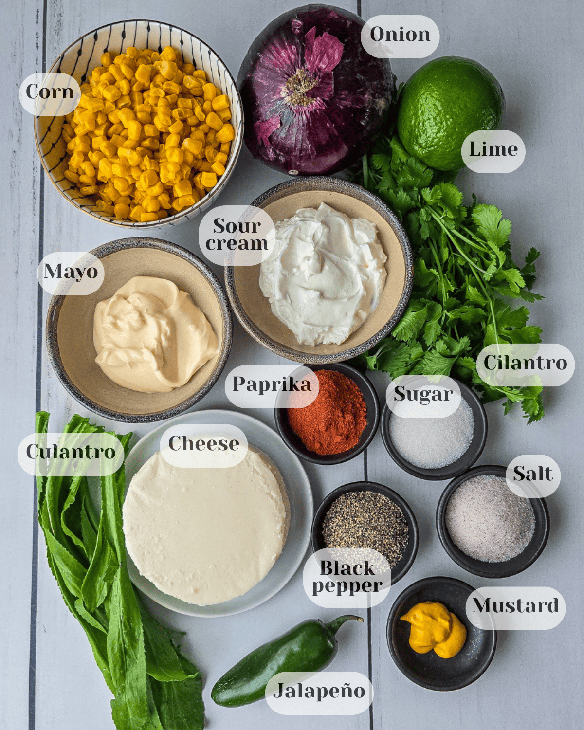 Photo taken from above with the ingredients for preparing the dip: a cup of sweet corn, a whole red onion, a whole lemon, a bunch of cilantro and culantro, a small bowl of mayonnaise, another one with sour cream, paprika, sugar, black pepper, salt, mustard, a small plate with round fresh cheese, and a jalapeño pepper.