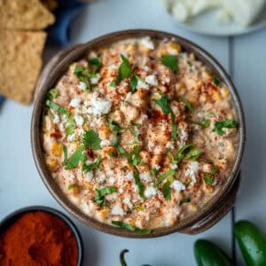 A deep bowl filled with corn dip, decorated on top with paprika, chopped cilantro, and cheese. The photo is taken from above; toasted tortilla chips and a small bowl of paprika can be seen on the side.