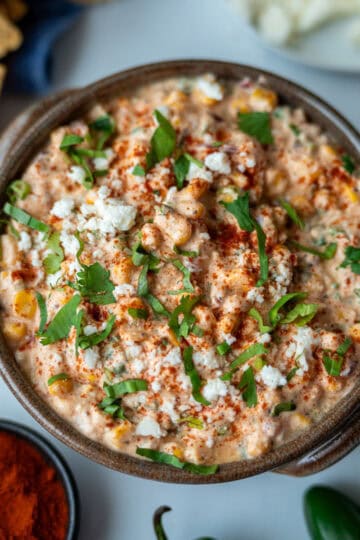 A deep bowl filled with corn dip, decorated on top with paprika, chopped cilantro, and cheese. The photo is taken from above; toasted tortilla chips and a small bowl of paprika can be seen on the side.