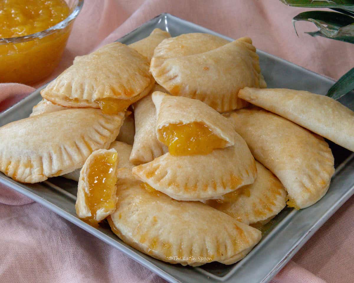 A square plate is holding pineapple empanadas on top. One of them is broken in half showing the pineapple filling