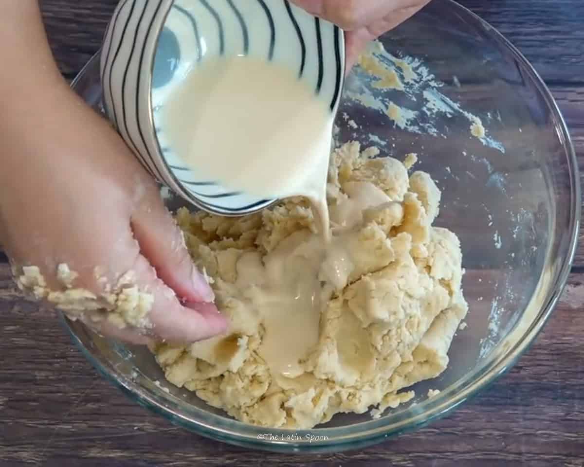 A glass bowl with a dough mixture being stirred by one hand is shown, while the other hand pours heavy cream from a cup.