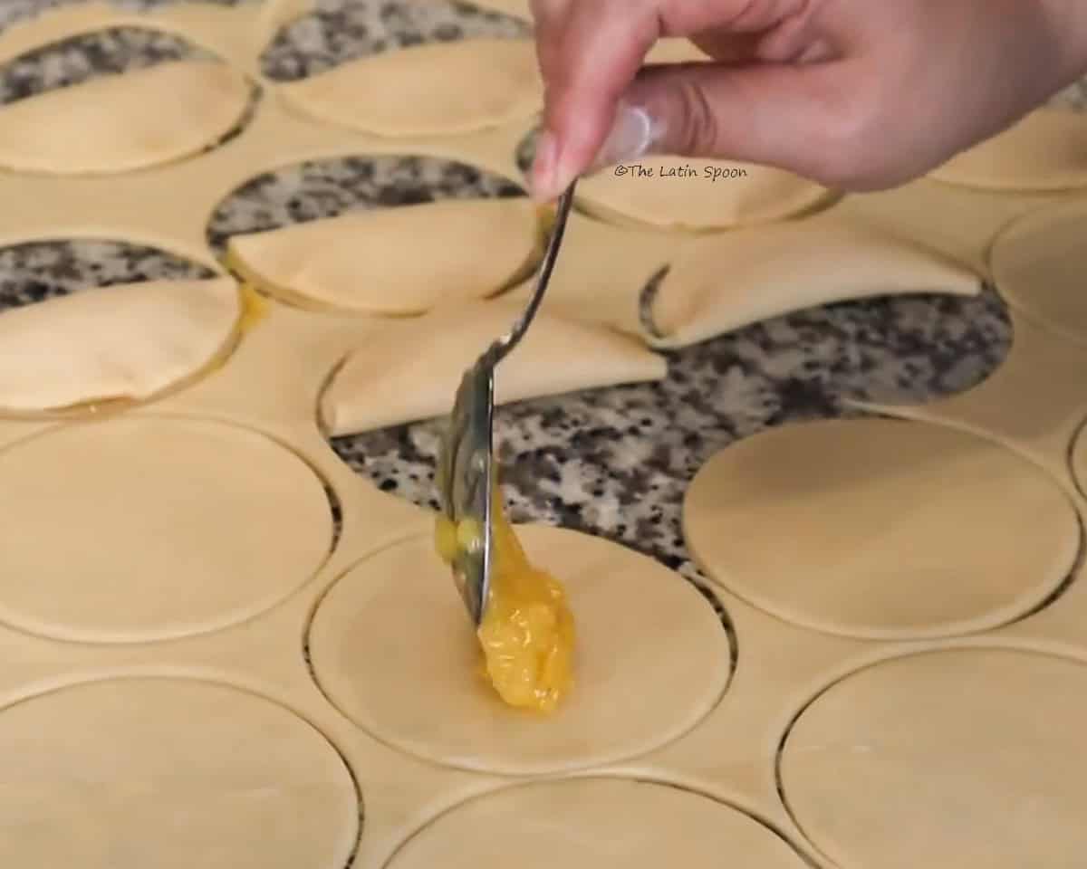 A fully rolled-out dough is shown on a countertop, cut into circles, while one hand holds a spoon and places pineapple filling in the center of one of the circles.