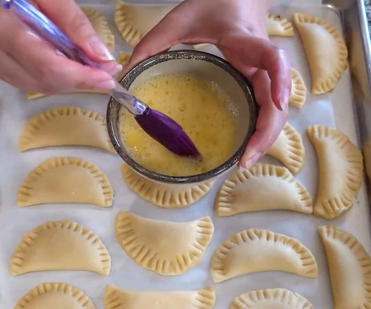 A tray of empanadas placed on parchment paper is shown. Two hands are holding a small cup with beaten egg and a pastry brush inside, ready to brush it over the empanadas.