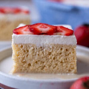A plate with a slice of tres leches cake, showing the fluffy sponge inside, topped with whipped cream and sliced strawberries.